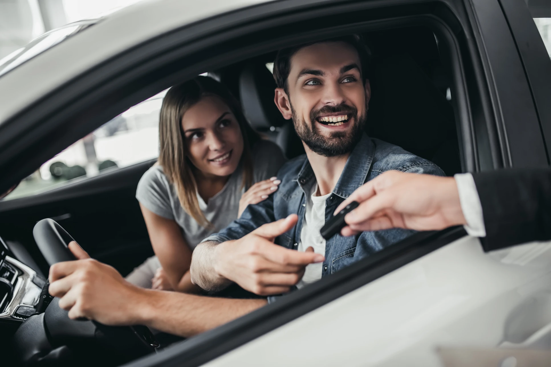 Homme souriant au volant d'une voiture moderne, femme passagère à ses côtés, tous deux regardent vers l'extérieur avec expression joyeuse