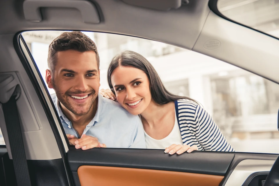 Couple souriant regardant par la fenêtre ouverte d'une berline blanche dans un environnement de concession automobile