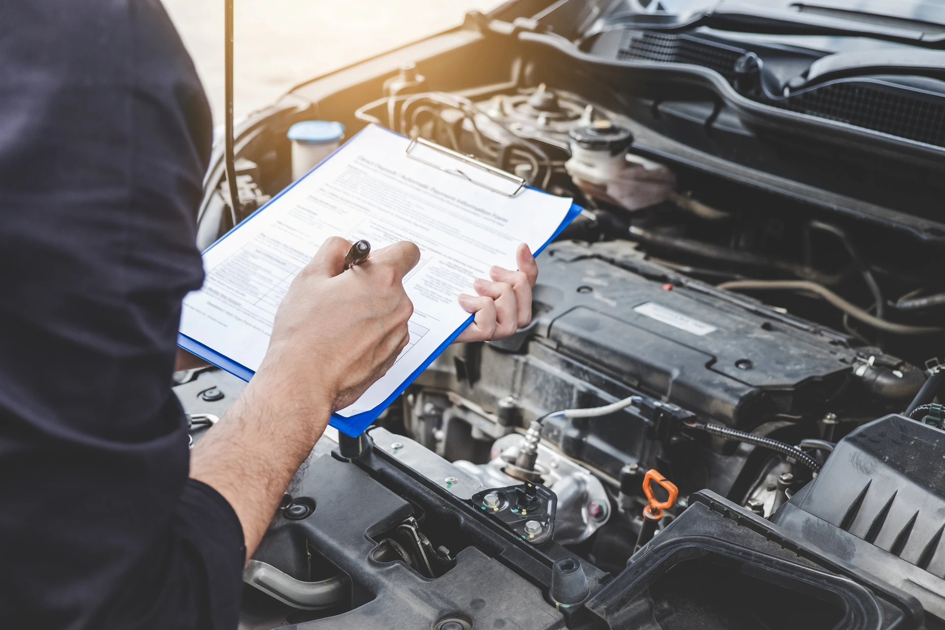 Mécanicien automobile inspectant un moteur de voiture avec capot ouvert, tenant un presse-papiers bleu avec formulaire de diagnostic