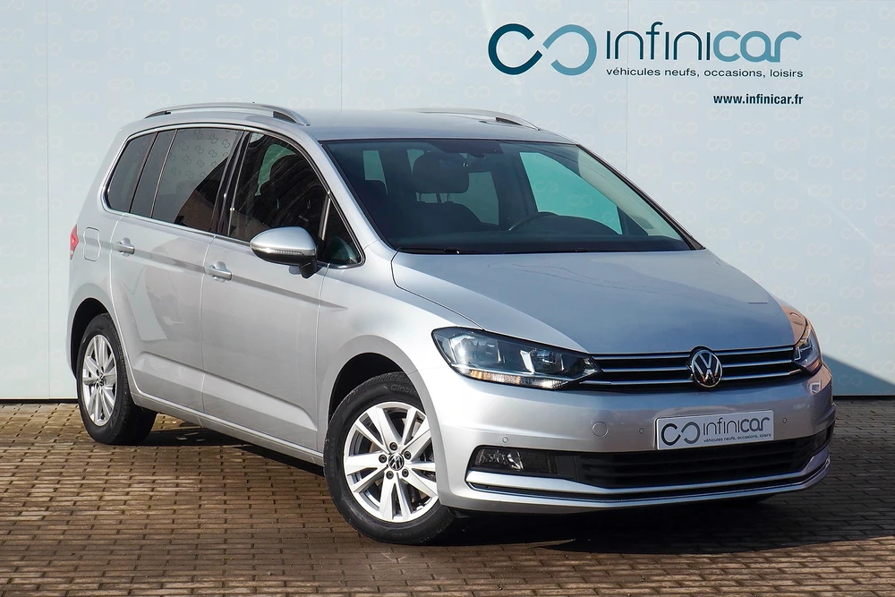 Monospace Volkswagen Touran de couleur gris argent, photographié en extérieur devant un fond publicitaire du concessionnaire Infinicar