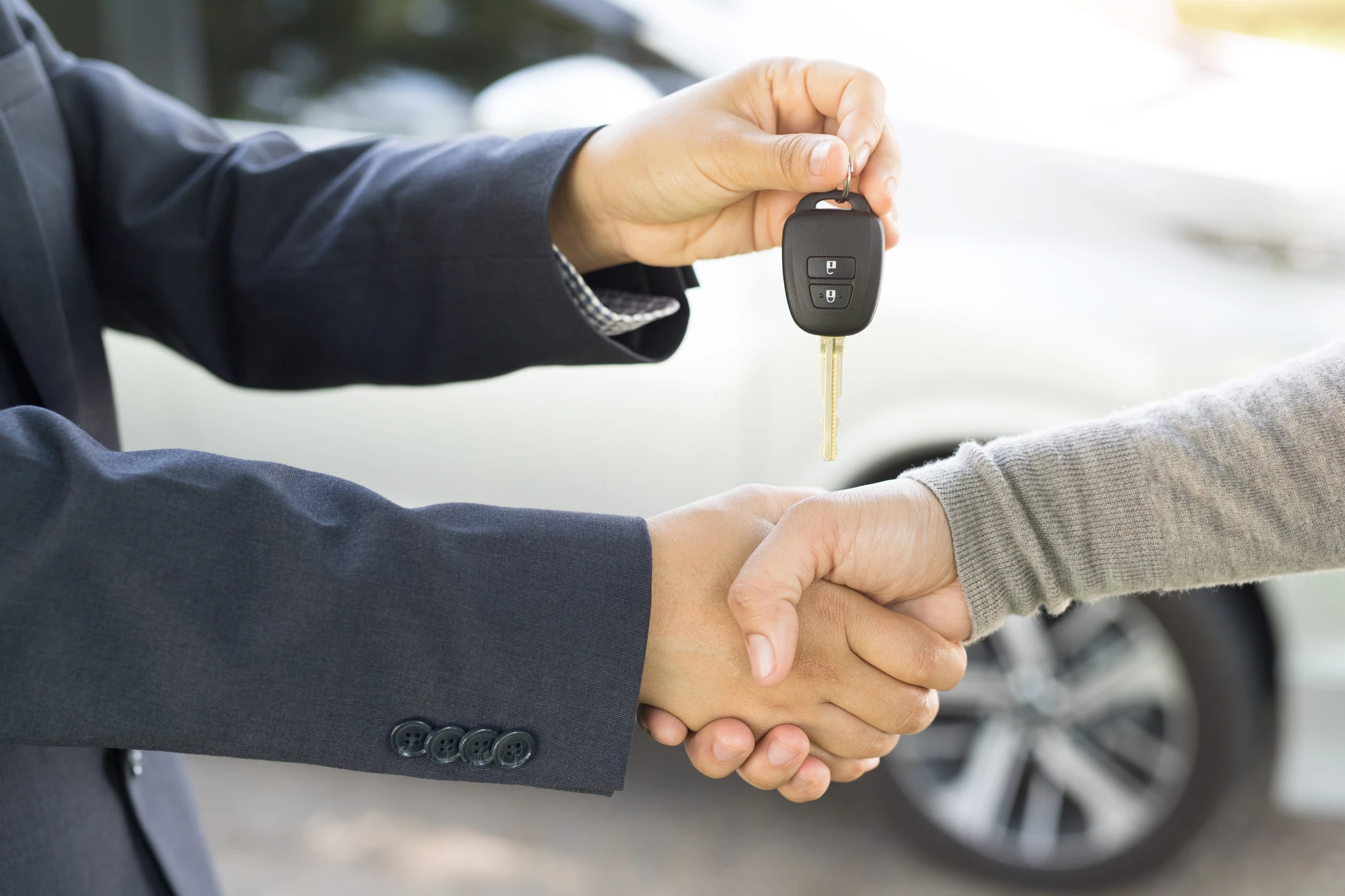 Deux personnes en costume effectuent une poignée de main professionnelle, l'une remettant des clés de voiture à l'autre dans un environnement automobile extérieur